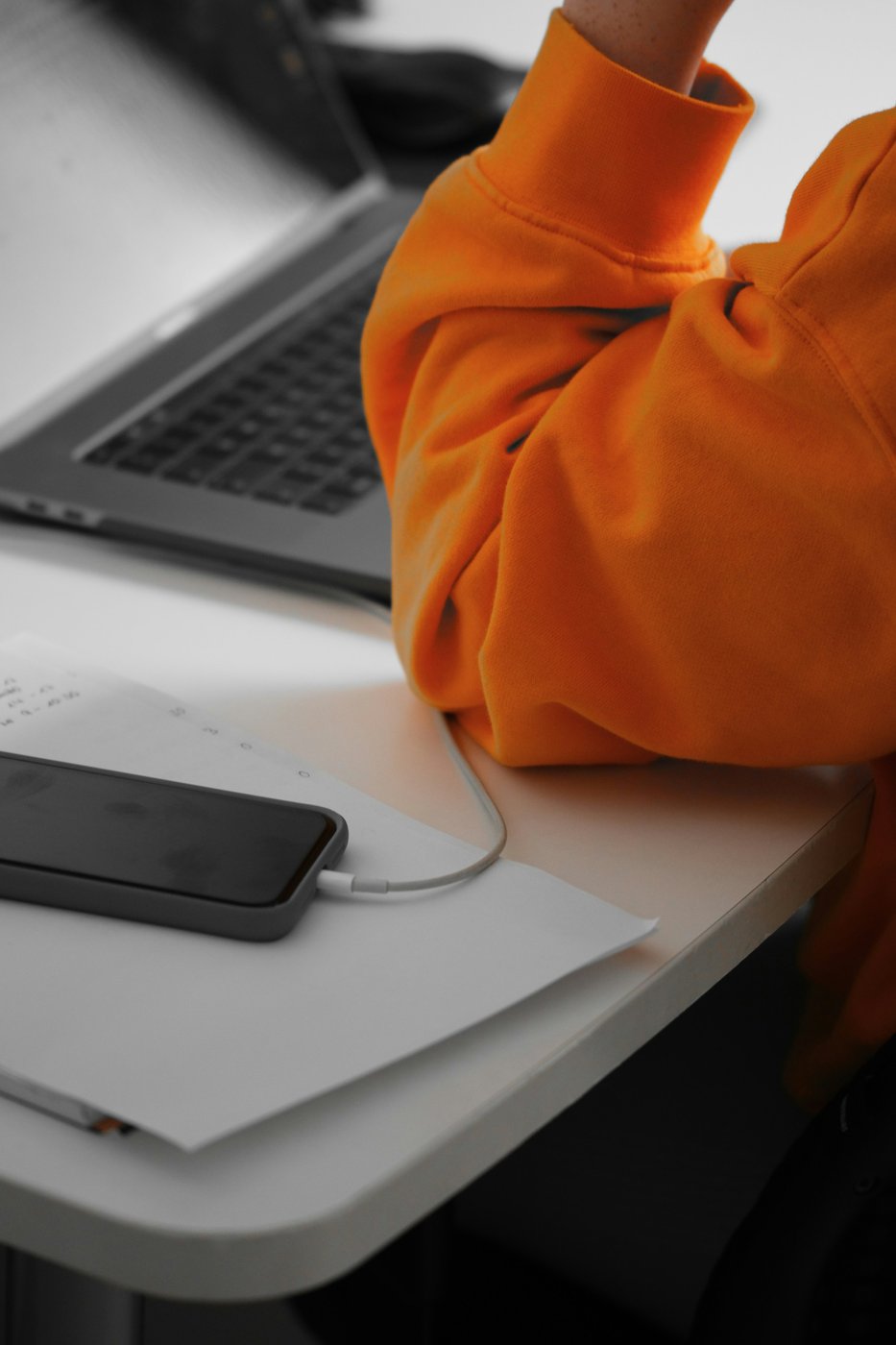 A person in an orange sweatshirt sits at a white desk beside a laptop, phone, and handwritten notes