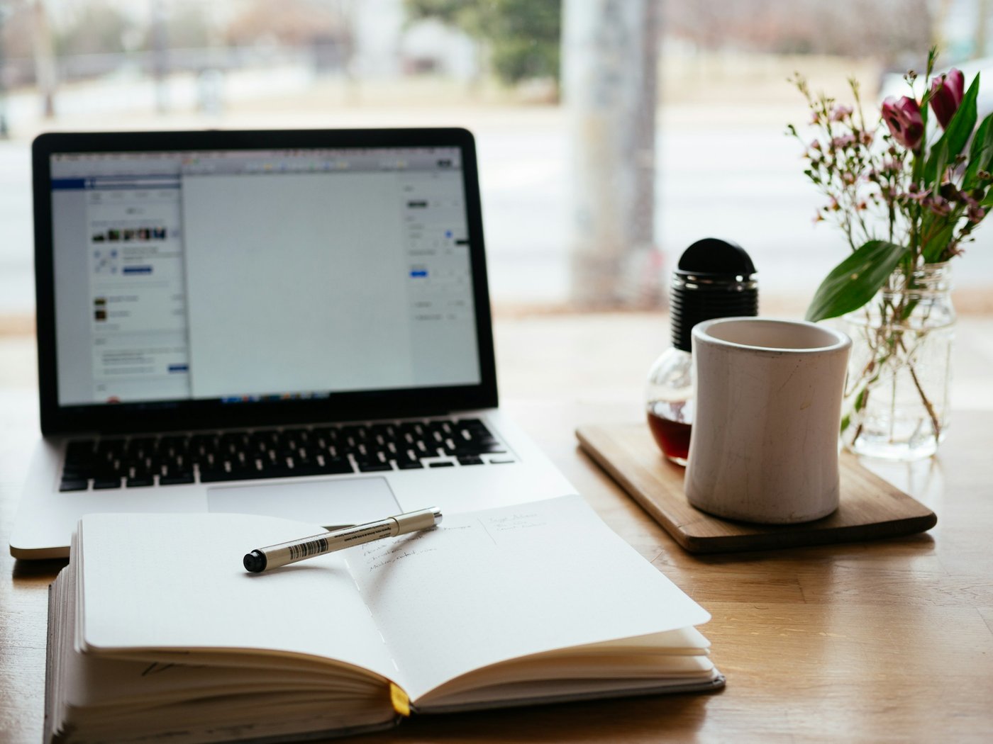 A laptop, open notebook, pen, and coffee sit on a wooden desk near a window
